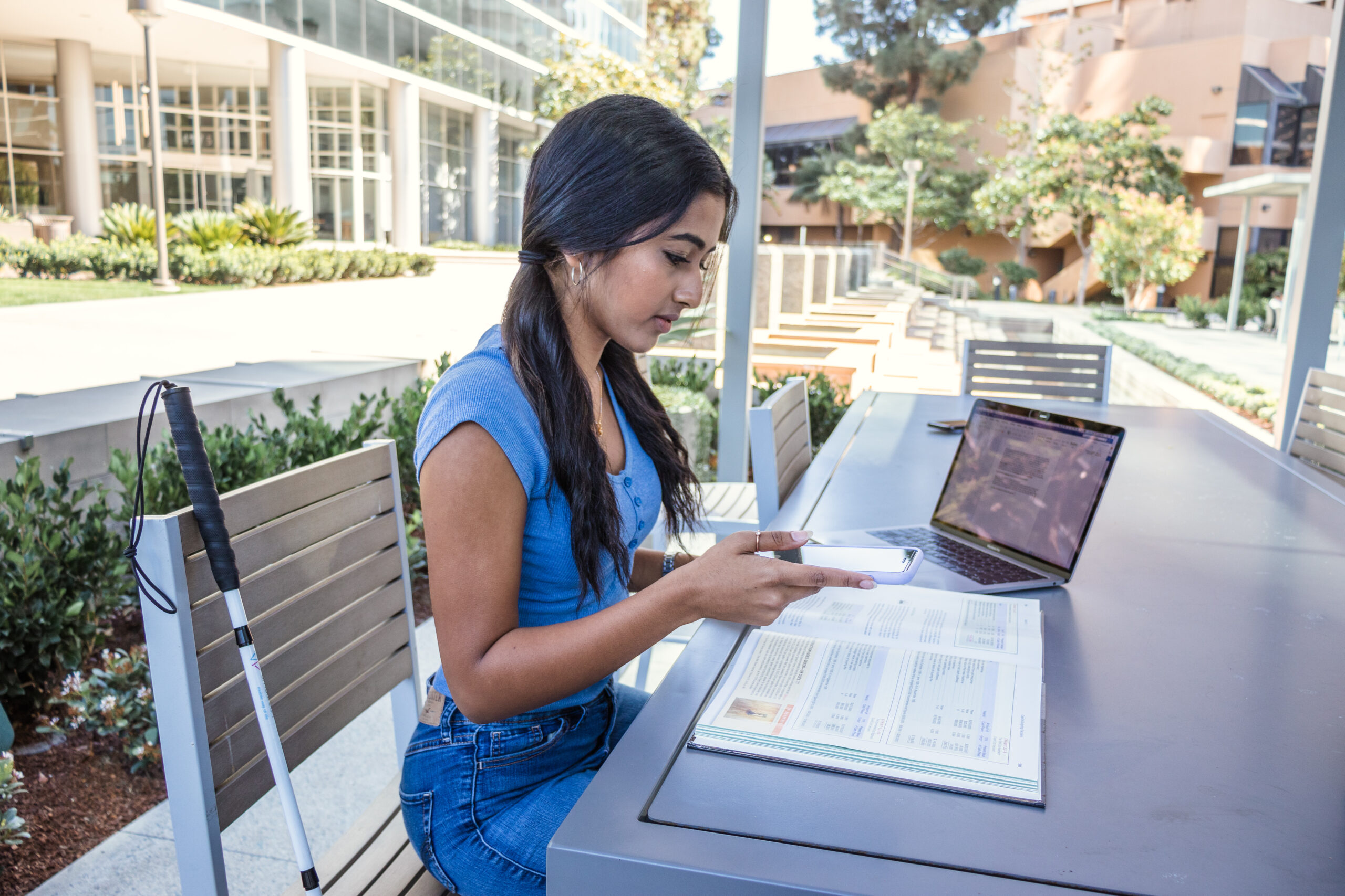 College student sitting at outdoor table using Aira to read non-Braille text book, has white cane by her side