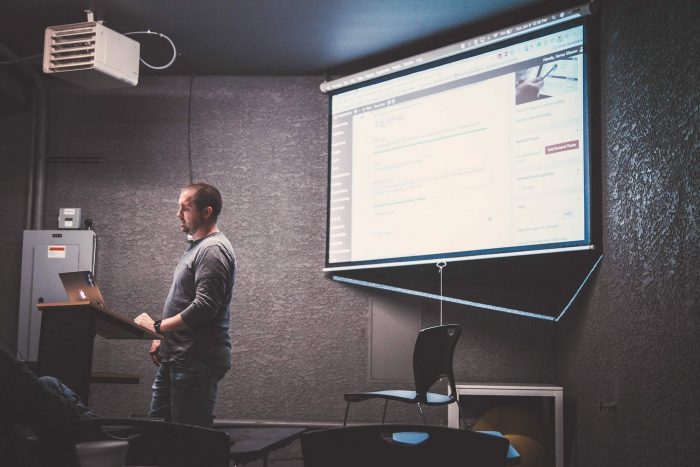 Classroom lecturer at podium, projecting laptop on screen behind