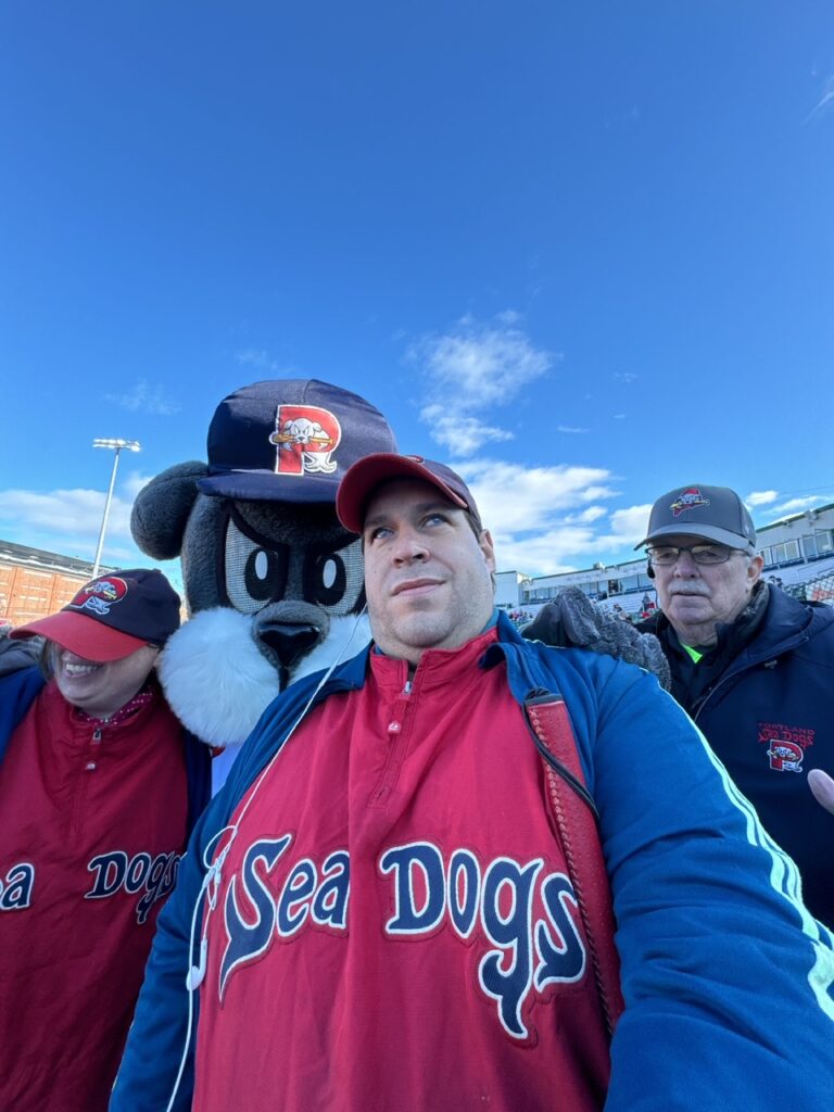 A blue sky and in the background and in the foreground, a selfie of several people. Randy Bellavance is in the centre, and on the left is Sarah Bellavance. In between them is Slugger, a life size dog mascot. Both Sarah and Randy wear Sea Dogs ball caps and red and blue jackets with "Sea Dogs" text on the front. 