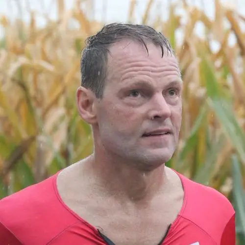A blurred field of yellow corn plants in the background, and athlete Dave Wilkinson standing in front of it wearing a red cycling jersey. He looks off camera to the right. 