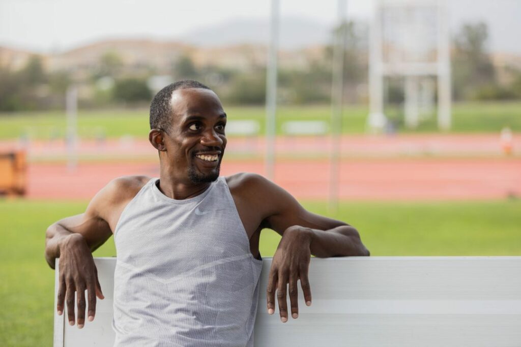A blurred background showing a green field with a clay track. Athlete Lex Gillette sits on the left hand side of a metal bench, smiling off to the right. Lex wears a grey running tank top and has his arms resting on the back of the bench. 