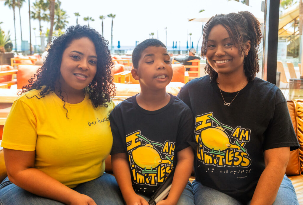 An outdoor scene with palm trees and plastic playground equipment in background. In foreground, Grayson Roberts sits next to two women, one at left wearing a yellow t-shirt. Grayson and the woman at right both wear black and yellow t-shirts with the words "I am limitless" on the front, with a large yellow lemon. 