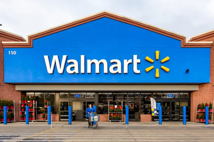 The front exterior of a Walmart location on a cloudy day. A woman in a blue shirt pushes a shopping cart, heading into the parking lot. 