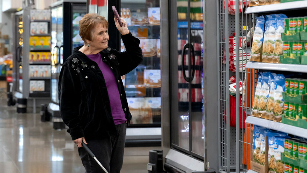 Paula Margeson, wearing a black cardigan and a purple shirt, walks through the freezer section of a Walmart. She holds a white cane in her right hand and with her left, holds a cellphone up to her ear. 