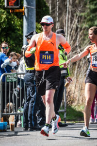 A man runs on the road during a marathon event. Spectators line the course. He wears an orange tank top, black shorts, a white hat, and running shoes. 