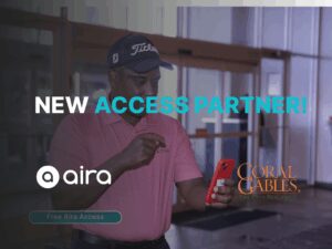 A man in a pink shirt holds up his phone and signs towards it. Text reads "New Access Partners." Aira and City of Coral Gables below.