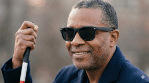 A man wearing black sunglasses and a navy blue jacket holds a cane with his right hand and smiles.